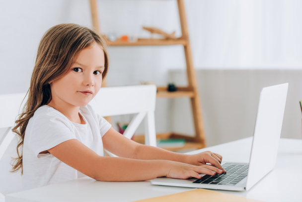 girl in white t-shirt looking at camera while sitting at table and using laptop  - Photo, image