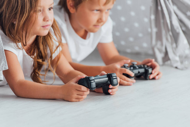 KYIV, UKRAINE - JULY 21, 2020: selective focus of brother and sister lying on floor and playing video game with joysticks - Fotografie, Obrázek