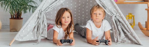 KYIV, UKRAINE - JULY 21, 2020: horizontal image of brother and sister in pajamas lying on floor in play tent and playing video game - Фото, изображение