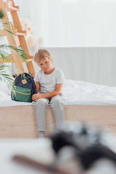Расстроенный мальчик в пижаме смотрит в камеру, сидя на кровати рядом со школьным рюкзаком  - Фото, изображение