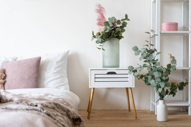 Hangulatos hálószoba apartman modern belsőépítészeti, virágok vázában éjjeliszekrény asztalon, kockás ágy felett és dekoráció a fa polcokon ellen fehér fal - Fotó, kép