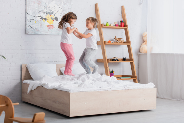 brat i siostra w piżamie trzymający się za ręce podczas skakania po łóżku - Zdjęcie, obraz