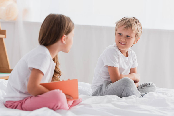 selective focus of girl with book looking at brother while sitting on bed in pajamas - Foto, Bild