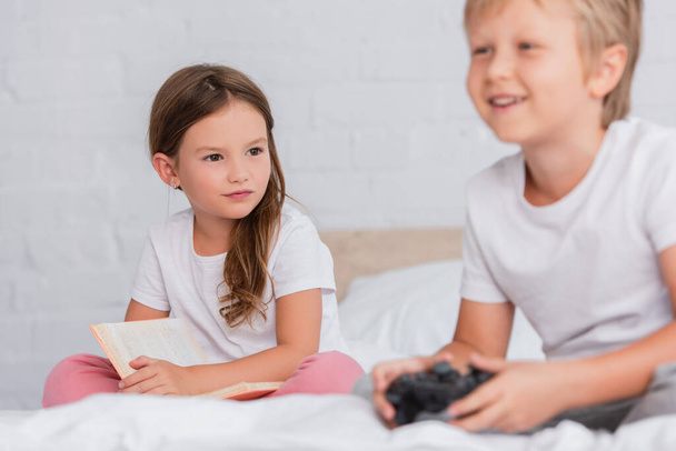 KYIV, UKRAINE - JULY 21, 2020: selective focus of girl with book looking at excited brother playing video game in bedroom - Fotografie, Obrázek