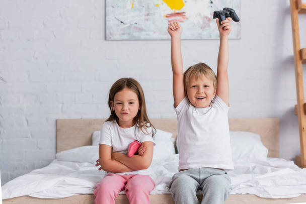 KYIV, UKRAINE - JULY 21, 2020: excited boy showing winner gesture while holding gamepad near offended sister - Photo, Image