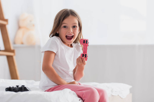 KYIV, UKRAINE - JULY 21, 2020: excited girl showing joystick while sitting on bed in pajamas - Фото, изображение