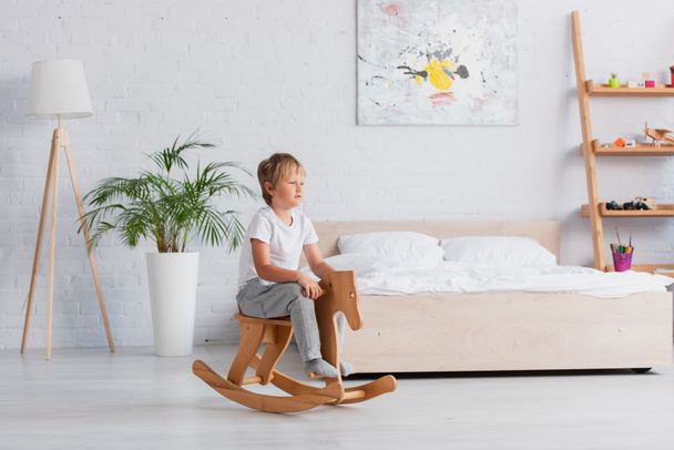 boy in pajamas riding rocking horse near potted plant and bed in spacious bedroom - Photo, Image