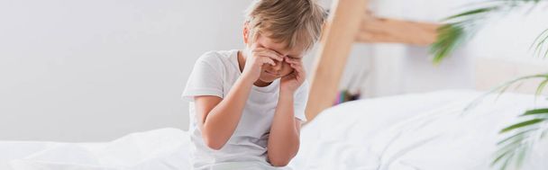 horizontaal beeld van ontwaakte jongen die ogen aanraakt terwijl hij in een pyjama op bed zit - Foto, afbeelding