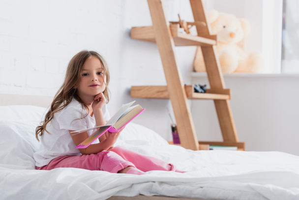 dziecko w piżamie patrząc w kamerę siedząc na łóżku i czytając książkę - Zdjęcie, obraz