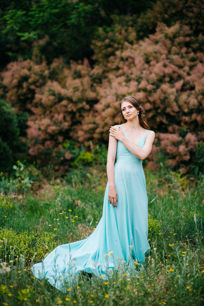 Happy girl in a turquoise long dress in a green park on a background of herbs, trees and rose bushes - Foto, Imagem