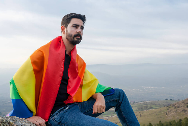 Mann schwul sitzend mit einer schwulen Flagge, die auf den Horizont blickt. Konzept von schwul, Stolz, Vielfalt - Foto, Bild