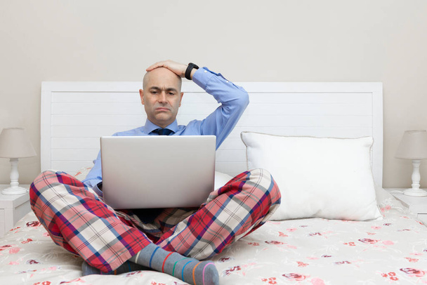 Homme travaillant avec un ordinateur portable sur un lit vêtu d'une chemise et d'une cravate et d'un pantalon de pyjama et la main sur la tête avec une expression inquiétante. Concept de télétravail - Photo, image