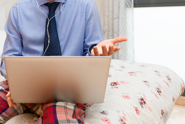 részlet a férfi dolgozik fejhallgató és egy laptop az ágyon öltözött ing és nyakkendő és pizsama nadrág egy kézzel magyarázza valamit a fejhallgató Teleworking koncepció - Fotó, kép