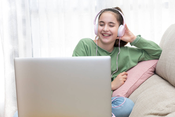 Fille assise sur un canapé regardant un ordinateur portable avec des écouteurs tout en souriant en jeans et un pull vert - Photo, image