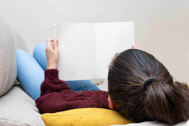 Menina com o cabelo amarrado deitado em um sofá com a cabeça em uma almofada lendo um livro
 - Foto, Imagem