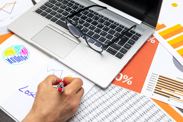 Χέρι ενός ατόμου που σχεδιάζει γραφικά σε ένα χαρτί με ένα φορητό υπολογιστή δίπλα του και γυαλιά σε ένα τραπέζι γεμάτο χαρτιά με αριθμούς και γραφικά - Φωτογραφία, εικόνα