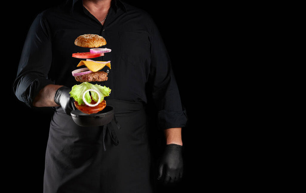adult man in a black uniform holding a cast iron round frying pan with frozen cheeseburger ingredients: sesame bun, cheese, tomato, onion, meat cutlet, black background, copy space - Foto, afbeelding