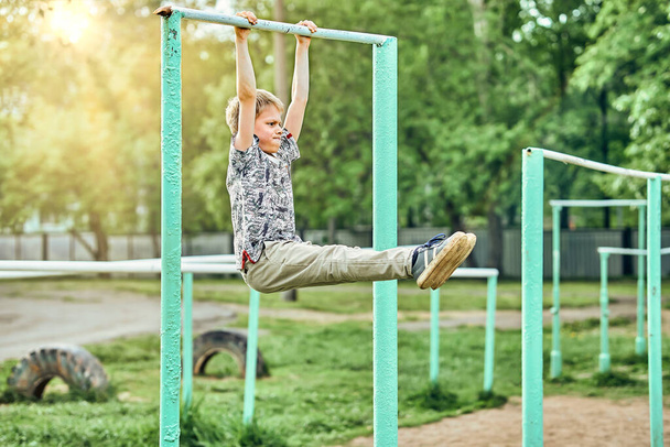 Full body side view of focused preteen boy in casual clothes doing exercises on metal bar while spending summer day on sports ground in park - Zdjęcie, obraz