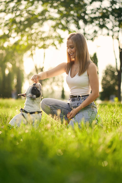 Jonge glimlachende vrouw training franse bulldog in park. - Foto, afbeelding