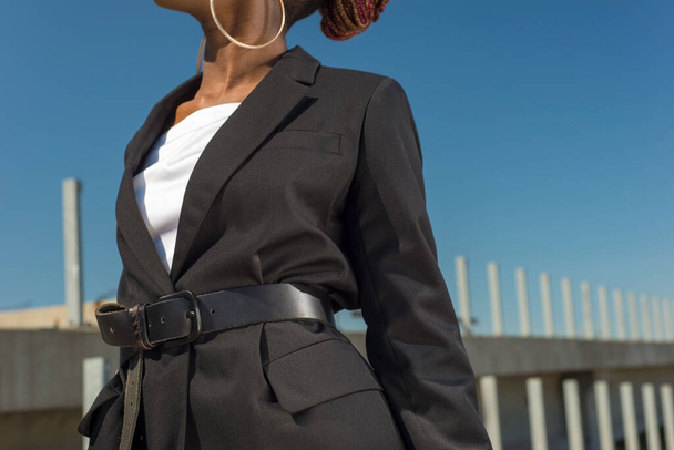 Nahaufnahme Details einer schwarzen Business-Jacke für Frauen, auf der Taille ein Ledergürtel eines afrikanischen Modells vor blauem Himmel. Stil, Design der Damenbekleidung. Schwarzes Mode-Mädchen. Afroamerikanisches Model zeigt eine neue Mode, die einen Gürtel trägt - Foto, Bild