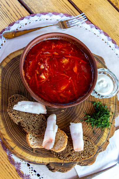 Свекольный суп в керамической тарелке с беконом, хлебом и сметаной на деревянной доске. Борх, традиционная украинская кухня - Фото, изображение