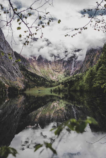 Baviera. Alemania. Cascada de Rothbach, montañas rocosas, bosque de pinos y cielo nuboso reflejan en el lago Obersee. Ramas de árboles y hojas en primer plano. Fondo de pantalla vertical - Foto, imagen