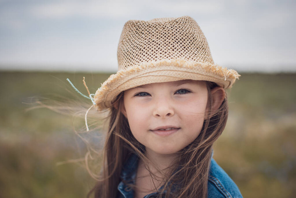 Portrait of a beautiful little girl in a straw hat and denim jacket against the backdrop of a rural road. - Foto, Imagem