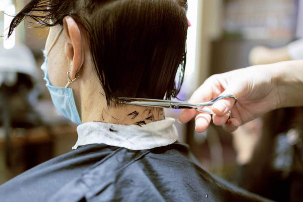 コロナウイルス中の美容サービス。フェイスマスクの美容師は顔のマスクで女性の髪をカットします。COVID-19中に髪を切る美容師 - 写真・画像