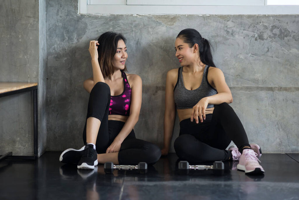 2 Beautiful fit athletic girls sit on floor of loft gym. Attractive women friends happy training and talk about sport exercise program with dumbbell equipment. bodybuilding and healthy lifestyle. - Photo, Image