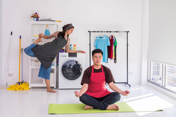 Önlüklü genç Asyalı karı koca çamaşır makinesini beklerken yoga yapıyorlar. Ev işleri ve hafta sonları temizlik.. - Fotoğraf, Görsel
