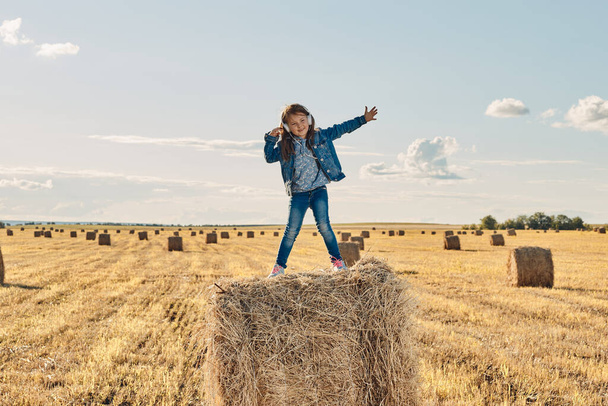 A cute little girl listens to music and dances on a haystack. - Photo, Image