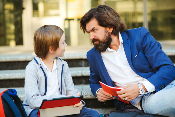 Father talking with son after lessons outside school. Schoolboy with backpack and notebooks. Dad and kid sitting at stairs before school. Cute son and fashionable dad having fun together. Fatherhood - Photo, Image
