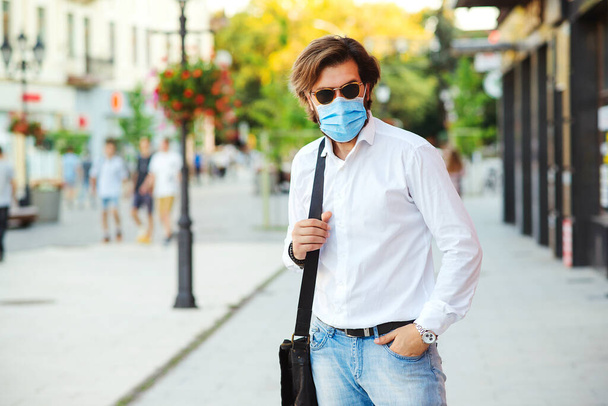 Γενειοφόρος με μάσκα ασφαλείας στο δρόμο. Ωραίος τύπος με ιατρική μάσκα μετά τη δουλειά. Μοντέρνος γενειοφόρος άντρας σε λευκό πουκάμισο με τσάντα. Μόδα, άνθρωποι και πραγματική ζωή. Καραντίνα από τον ιό Coronavirus. - Φωτογραφία, εικόνα