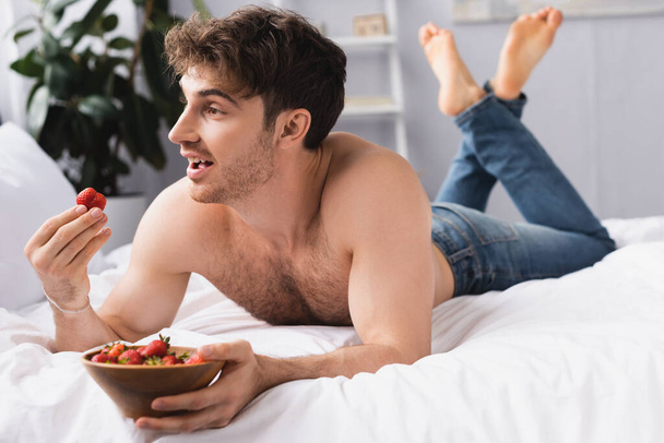 selective focus of shirtless and barefoot man lying on bed and holding bowl with ripe strawberries  - Photo, Image