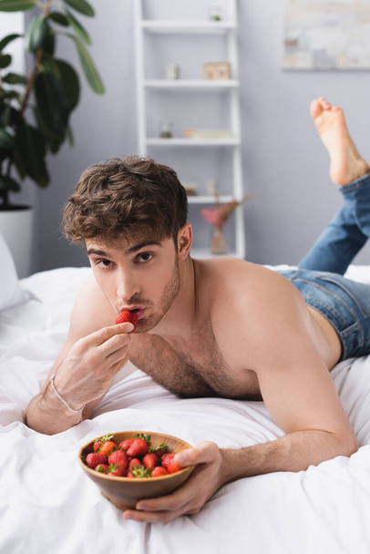 enfoque selectivo del hombre sin camisa acostado en la cama y sosteniendo tazón con fresas maduras  - Foto, imagen