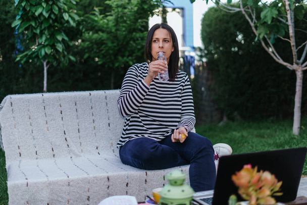 Teleworking. Woman drinking water while is sitting in the garden dressing white and blue stripes jersey and blue jeans, having on the table a laptop, cellphone and infusion. - Photo, Image