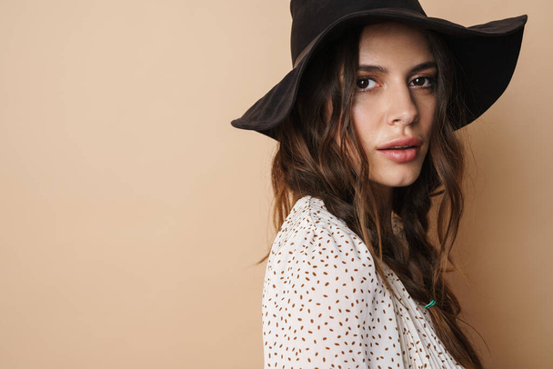 Photo of brunette beautiful woman in hat posing and looking at camera isolated over beige background - Photo, Image
