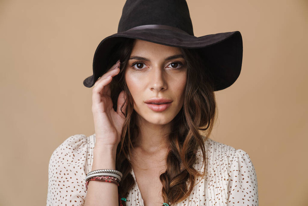 Photo of brunette beautiful woman in hat posing and looking at camera isolated over beige background - Fotó, kép
