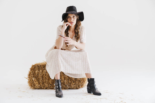 Photo of brunette seductive woman looking at camera while sitting on hay isolated over white background - Photo, Image