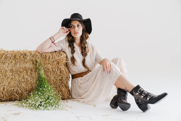 Photo of attractive young woman wearing stylish hat posing with flowers and hay isolated over white wall - 写真・画像