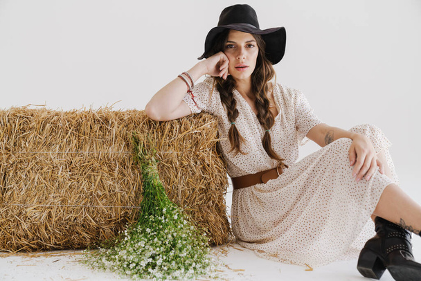 Photo of attractive young woman wearing stylish hat posing with flowers and hay isolated over white wall - Foto, Imagen