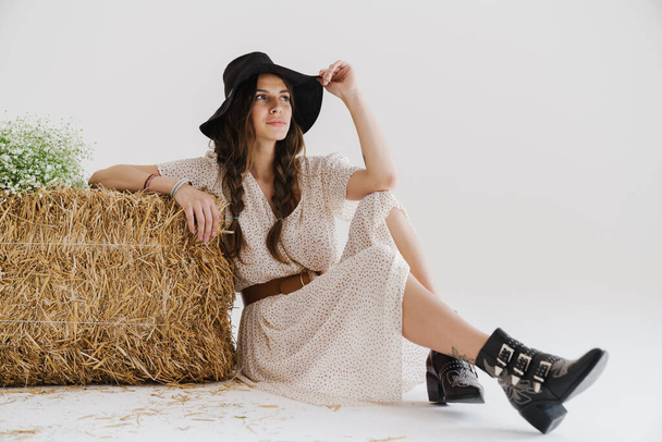 Photo of pleased young woman wearing stylish hat posing with hay while sitting on floor isolated over white wall - Photo, image