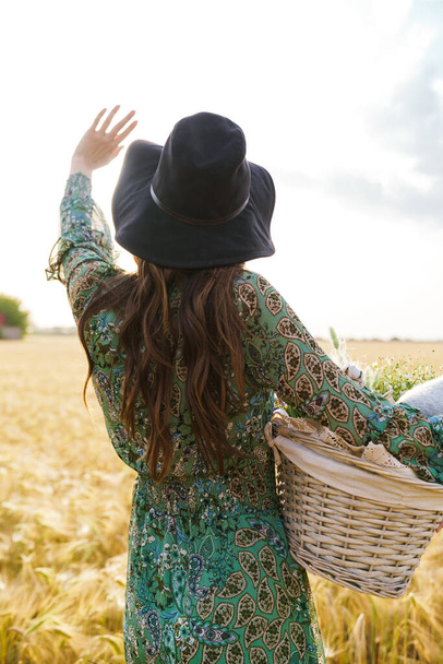 Photo from back of cute woman in stylish hat waving hand while walking with basket at picnic on wheat field - Foto, Imagen