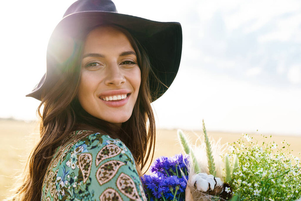 Photo of happy young woman wearing stylish hat smiling while walking with flowers at picnic on wheat field - Foto, Imagen