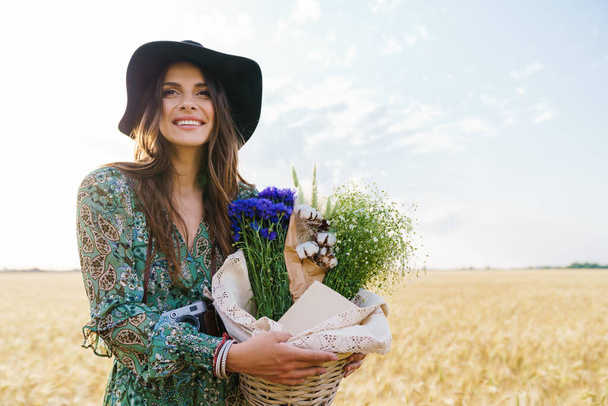 Photo of happy young woman wearing stylish hat smiling while walking with basket at picnic on wheat field - Fotoğraf, Görsel
