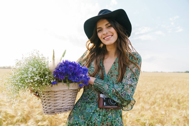 Photo of happy young woman wearing stylish hat smiling while walking with basket at picnic on wheat field - Fotografie, Obrázek