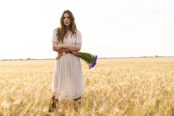 Photo of beautiful cute woman in stylish dress posing while walking with flowers on wheat field - Foto, afbeelding