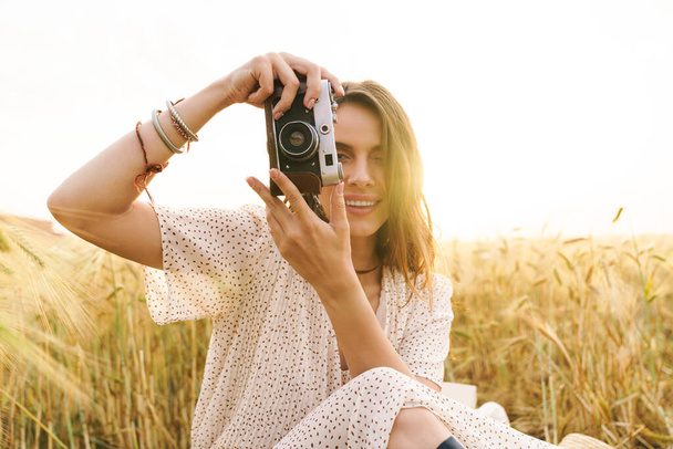 Photo of charming joyful woman in stylish dress using retro camera while sitting on wheat field - Fotó, kép