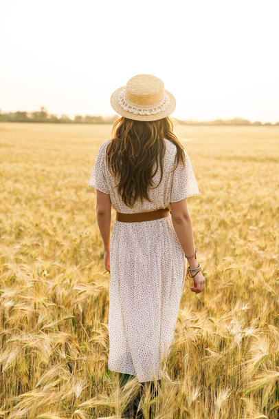 Photo from back of cute brunette woman in straw hat walking on wheat field at summer day - Фото, изображение