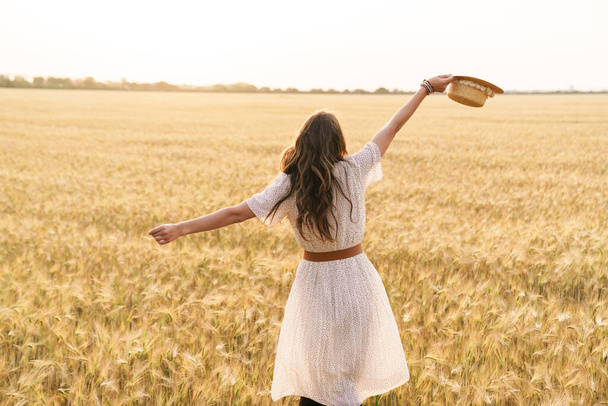Photo from back of cute brunette woman with straw hat dancing on wheat field at summer day - Foto, Imagen